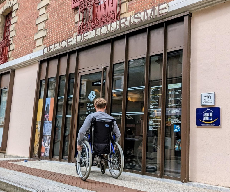 Un jeune homme de dos en fauteuil roulant s'apprête à entrer dans un office de tourisme