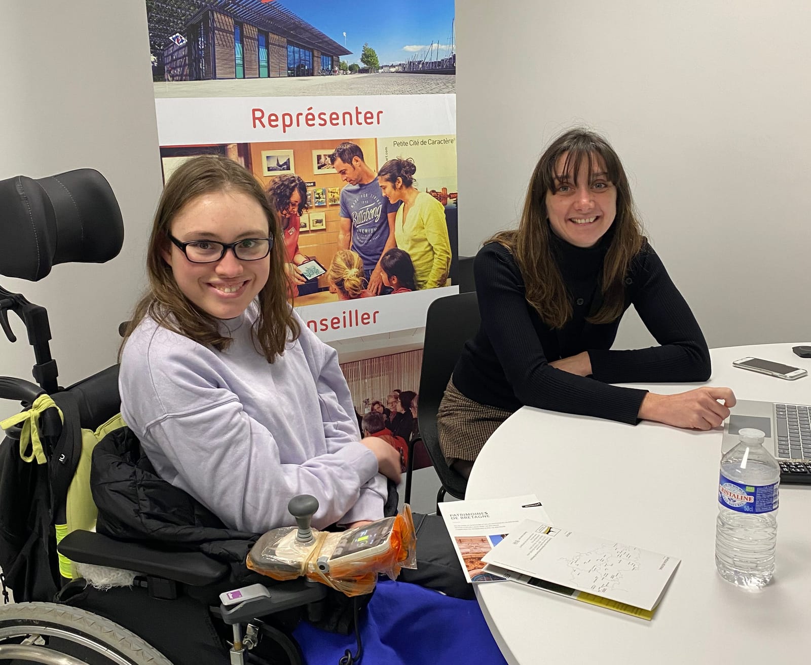 Un perosnne en situation de handicap moteur est assise à côté d'une autre personne autour d'une table, derrière un kakémono i